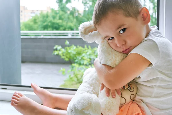 Niño de dos años sentado junto a la ventana y abraza a un conejito de juguete. tiempo lluvioso, esperando a que papá vuelva a casa del trabajo —  Fotos de Stock