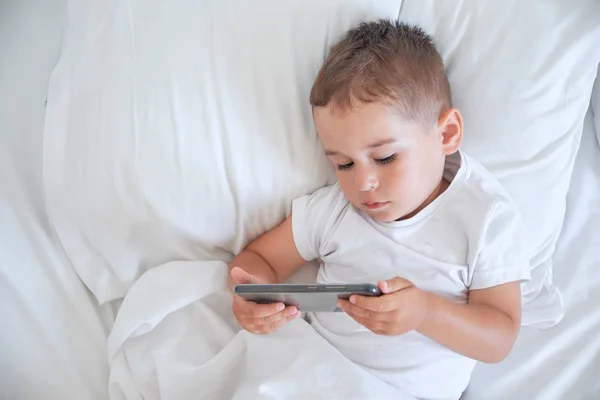 Kisfiú lefekvés és néz rajzfilmeket a smartphone előtt — Stock Fotó