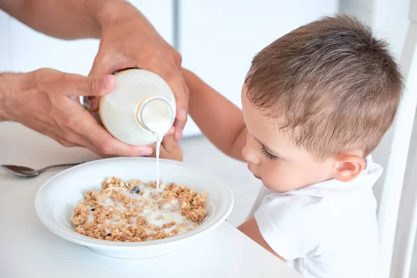 O rapazinho ajuda o pai a deitar leite em farinha de aveia. Pequeno-almoço rápido em casa — Fotografia de Stock
