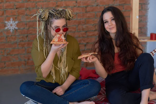 two girls eat pizza in the room on the bed. junk food.