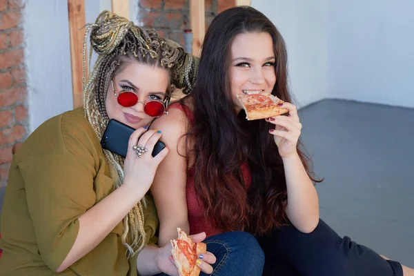 two girls eat pizza in the room on the bed. junk food.