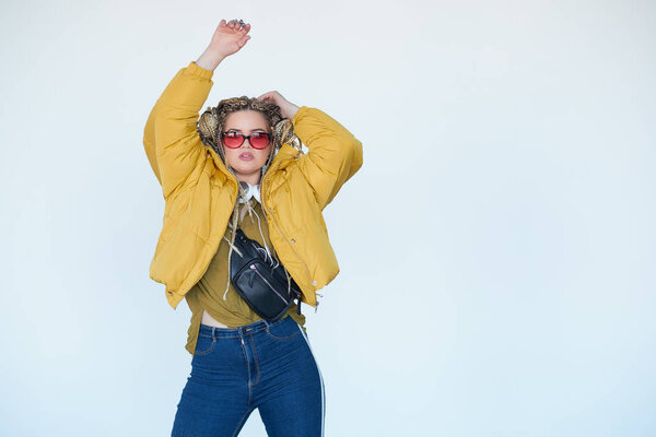 young blonde girl in sunglasses with african pigtails listens to music in headphones and dances against a white background.