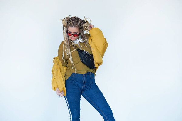 young blonde girl in sunglasses with african pigtails listens to music in headphones and dances against a white background.