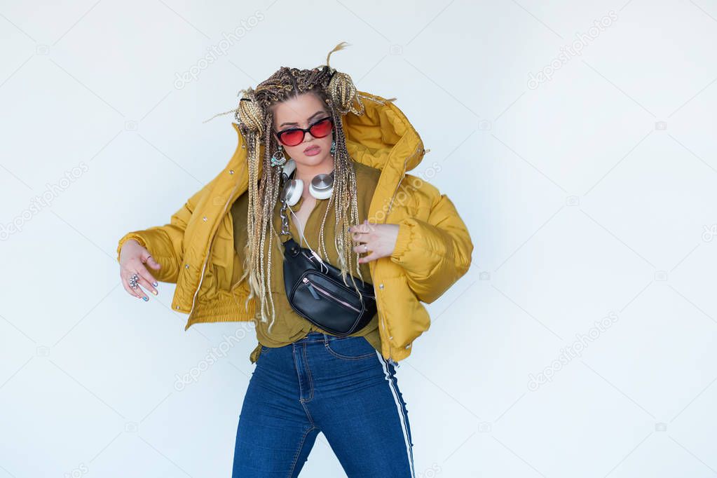 young blonde girl in sunglasses with african pigtails listens to music in headphones and dances against a white background.