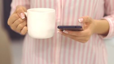 Kadın sıcak çay içiyor ve cep telefonu ekranında bakmak. Çekim WOMANS eller.