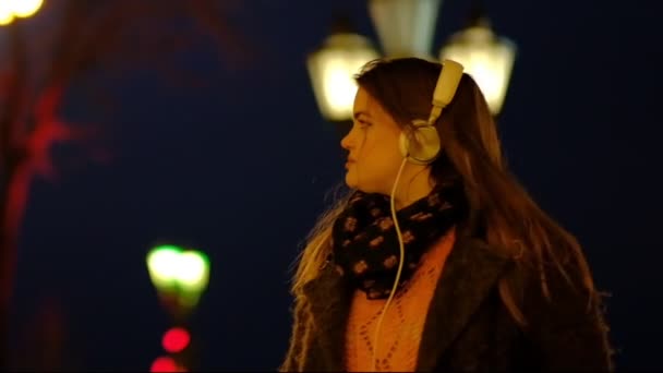 Muchacha adolescente de primer plano con el pelo largo caminando por la calle, escuchando la música con auriculares en la noche bajo luces rojas. En cámara lenta. . — Vídeo de stock