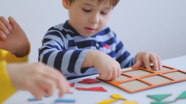 Menino asiático pouco coleta quebra-cabeças com a mãe sentada na mesa. Uma criança bonita recolhe uma imagem de grandes detalhes multi-coloridos. A brincar com um puzzle. câmara lenta. Professor brincando com menino — Vídeo de Stock