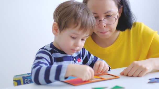 Piccolo ragazzo asiatico raccoglie puzzle con mamma seduta alla scrivania. Un bambino carino raccoglie un'immagine da grandi dettagli multicolori. Giocare con un puzzle. Al rallentatore. Insegnante giocare con ragazzo — Video Stock