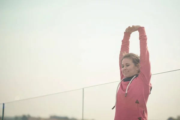 Mujer joven en un traje rosa se está preparando para el entrenamiento deportivo o la carrera atlética, entrenamiento de la mañana. madrugada de verano en el centro de la ciudad. vista desde las multitudes laterales en la distancia . — Foto de Stock