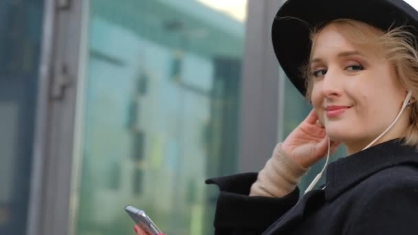 Agradable chica rubia en abrigo negro y sombrero escucha música con auriculares para teléfonos inteligentes contra la calle de la ciudad cámara lenta — Vídeo de stock