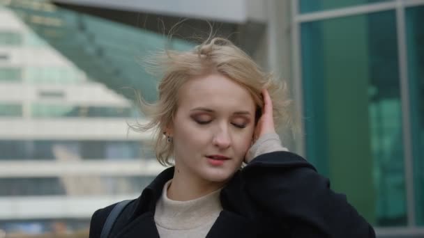 Atractivo rubia chica en negro abrigo poses para cámara contra borrosa moderno edificio cerrar vista cámara lenta . — Vídeos de Stock