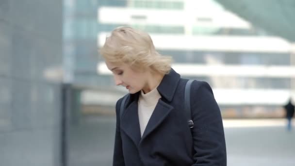 Atractivo rubia chica en negro abrigo poses para cámara contra borrosa moderno edificio cerrar vista cámara lenta — Vídeos de Stock