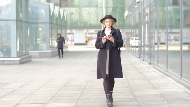 Chica en negro textos en el teléfono caminando a lo largo de cristal ventana pared — Vídeos de Stock