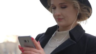 siyah ceket ve şapka güzel sarışın kız şehir sokak yavaş hareket karşı akıllı telefon kulaklıkile müzik dinler