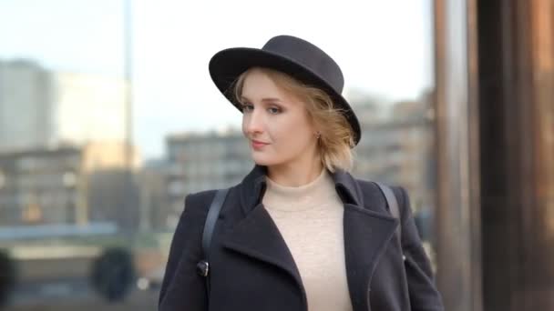 Close pretty girl in black coat holds hat listening to music with headphones against blurry modern building slow motion — Stock Video