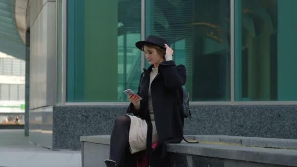 Agradable chica rubia en abrigo negro y sombrero escucha música con auriculares para teléfonos inteligentes contra la calle de la ciudad cámara lenta — Vídeos de Stock