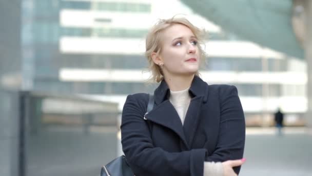 Attractive blonde girl in black coat poses for camera against blurred modern building close view slow motion — Stock Video