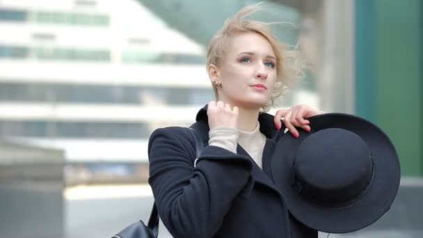Attraente ragazza bionda in cappotto nero posa per la fotocamera contro offuscata edificio moderno vista da vicino rallentatore — Video Stock