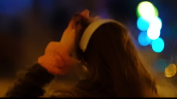 Zbliżenie nastoletnie dziewczyny z długimi włosami chodzenie wzdłuż ulicy, słuchanie muzyki noszenie słuchawek w nocy pod czerwonym światłem. W zwolnionym tempie. — Wideo stockowe