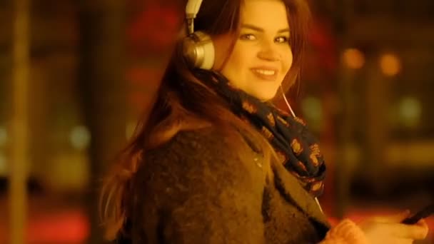 Closeup teenage girl with long hair walking along the street,listening to the music wearing headphones at night under red lights. Slow-motion. — Stock Video