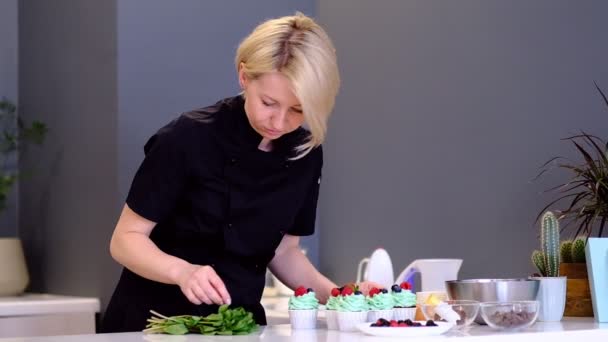 Postre cocinero mano femenina decora sabrosos cupcakes crema de turquesa cubierta con frambuesas y arándanos, menta hoja cocina — Vídeo de stock