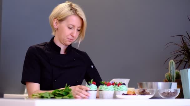 Dessertköchin weibliche Hand dekoriert leckere Cupcakes Türkis Sahnebelag mit Himbeeren und Blaubeeren, Minzblätter in der Küche — Stockvideo