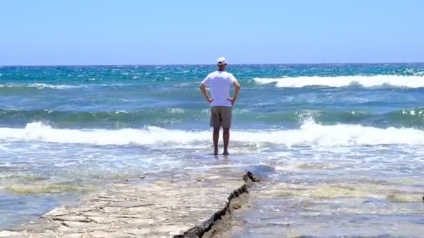 Человек стоит на пляже и наблюдает за волнами. Турист любуется прекрасным видом на дикий пляж и дышит свежим морским воздухом — стоковое видео
