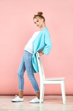 Güzel sarışın kız pembe bir arka plan üzerinde beyaz bir sandalye yanında duruyor. tarzı genç kız. Stüdyo çekimi.