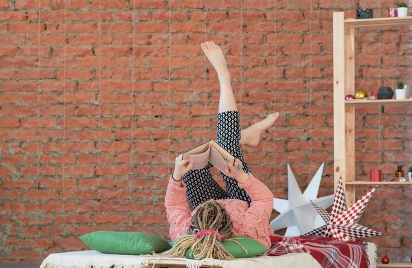 Jeune femme lisant le livre sur le lit à la maison. Un mur de briques. loft. week-end paresseux . — Photo