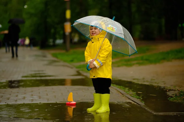 Bambino felice sta giocando in una pozzanghera in un giorno piovoso estivo. un ragazzo con un impermeabile giallo cammina nel parco . — Foto Stock