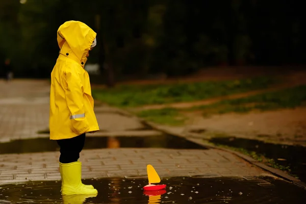 Boy in a yellow raincoat looks at a toy ship that floats in a puddle. little baby walking in the park. — Stock Photo, Image