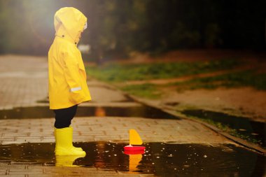 sarı yağmurluk lu çocuk su birikintisinde yüzen bir oyuncak gemiye bakar. parkta yürüyen küçük bebek.