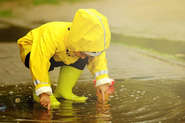 Bambino carino che gioca in una pozzanghera con una nave in un giorno di pioggia estivo. un ragazzo con un impermeabile giallo cammina nel parco . — Foto Stock