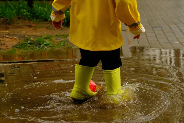 child jumps in a puddle arranges a storm for a small plastic boat. legs in yellow rubber boots.