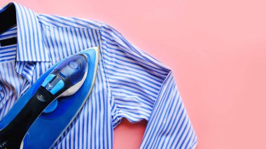 Pembe arka plandaki ütü masasında elektrikli ütü ve gömlek. Demir mavisi çizgili gömlek.