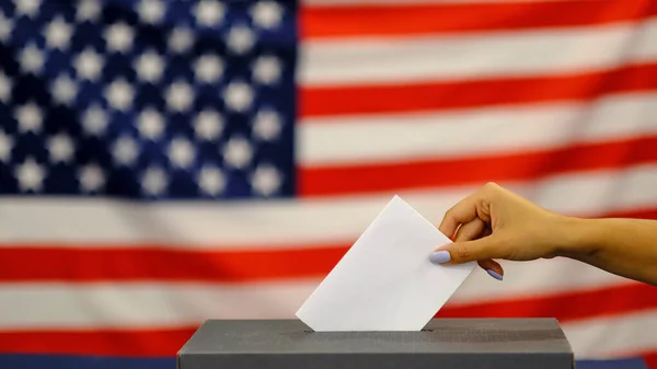 woman putting a ballot in a ballot box on election day. Close up of hand with white votes paper on usa flag background.