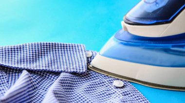 Pembe arka plandaki ütü masasında elektrikli ütü ve gömlek. Demir mavisi çizgili gömlek.