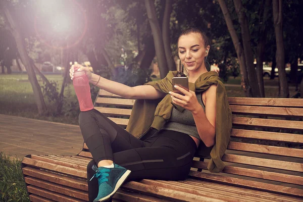 Beautiful girl athlete summer city, resting after fitness workout. Sportswear leggings top. Shaker with water protein skipping, rope smartphone headphones. Free space. Listening music. — Stock Photo, Image