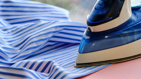 Electric iron and shirt on ironing board in room on a pink background. iron blue striped shirt — Stock Photo, Image