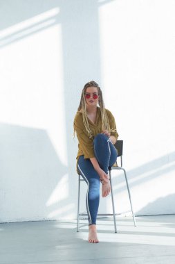 Yuvarlak güneş gözlüklü sevimli genç bayan stüdyoda bir sandalyede oturuyor. Beyaz bir duvara yaslan.