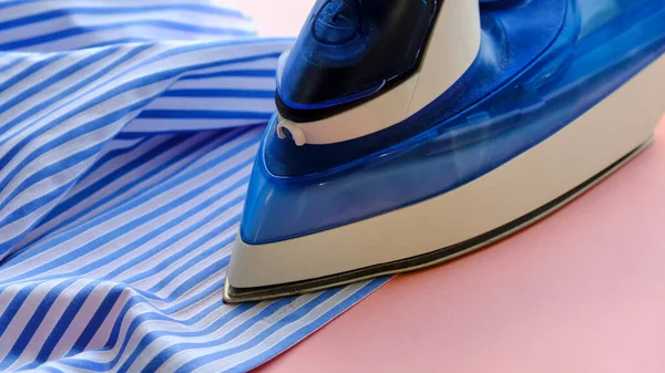 Electric iron and shirt on ironing board in room on a pink background. iron blue striped shirt — Stock Photo, Image