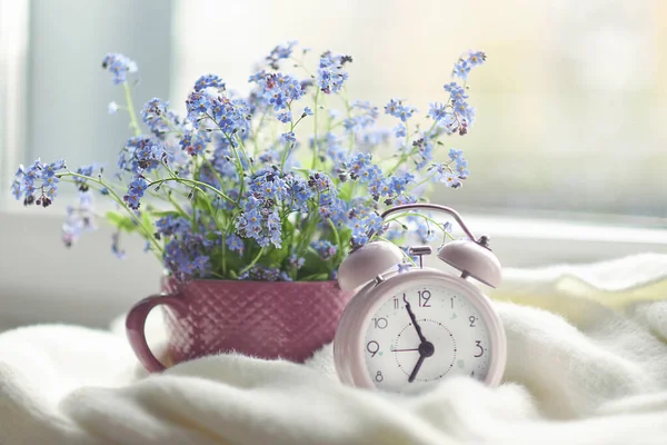 Bir buket çiçek ve pencere pervazında rahat beyaz bir ekose. Pencerenin yanındaki pembe saat sabah saatini gösteriyor. — Stok fotoğraf