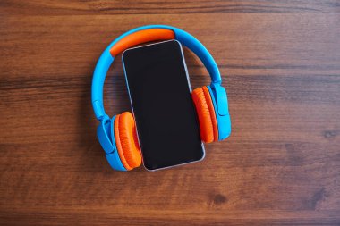 Ahşap bir masada boş ekranlı ve kulaklıklı akıllı telefon. Müzik ve teknoloji konseptinin en üst noktası. Kulaklıklı, düz akıllı telefon. Yaratıcı tasarım metni ve sözcük için kopyalama alanı.