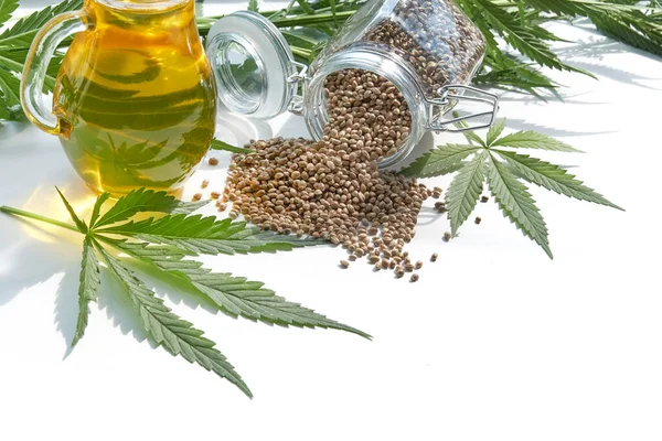 テーブルの上の大麻の種、ガラス瓶の油、白い背景に隔離された大麻の葉 — ストック写真