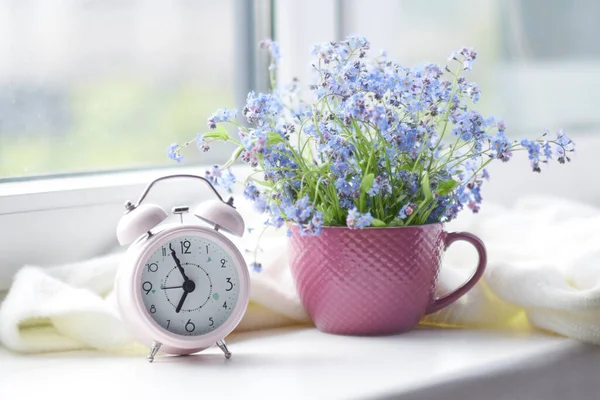 Spring bouquet of flowers and a cozy white plaid on the windowsill. pink clock by the window shows morning time — Stock Photo, Image