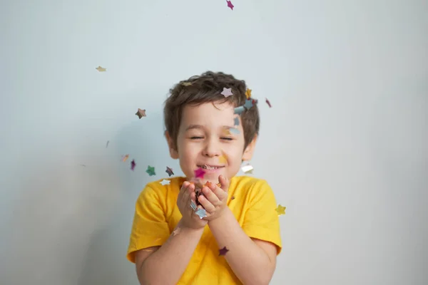 생일 축하 해. 매력적 이고 귀엽고 귀여운 작은 소년이 감정적 인 표정으로 축제 분위기를 보여 주기 위해 콘 페티를 불고 있는 사진. — 스톡 사진