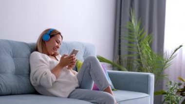 Yakından bakınca Asyalı kaygısız genç bir kadın kanepeye yaslanmış akıllı bir telefon tutuyor internette sohbet ediyor kablosuz kulaklıkla müzik dinliyor şarkı söylüyor, boş zaman konsepti.