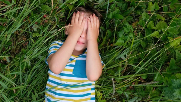 A fiú a zöld fű felett rejtegeti a szemét. Egy kisfiú, aki behunyja a szemét, kezével és tenyerével nevetve sikoltozik.. — Stock Fotó