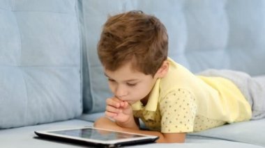 Anaokulu çocuğu evdeki kanepede otururken online eğitim için tablet bilgisayar kullanıyor. Çocuk internetten okumak için başvuru kullanıyor. Aleti olan çocuk.