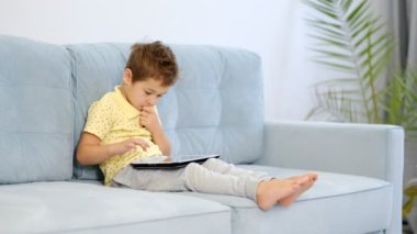 Anaokulu çocuğu evdeki kanepede otururken online eğitim için tablet bilgisayar kullanıyor. Çocuk internetten okumak için başvuru kullanıyor. Aleti olan çocuk.